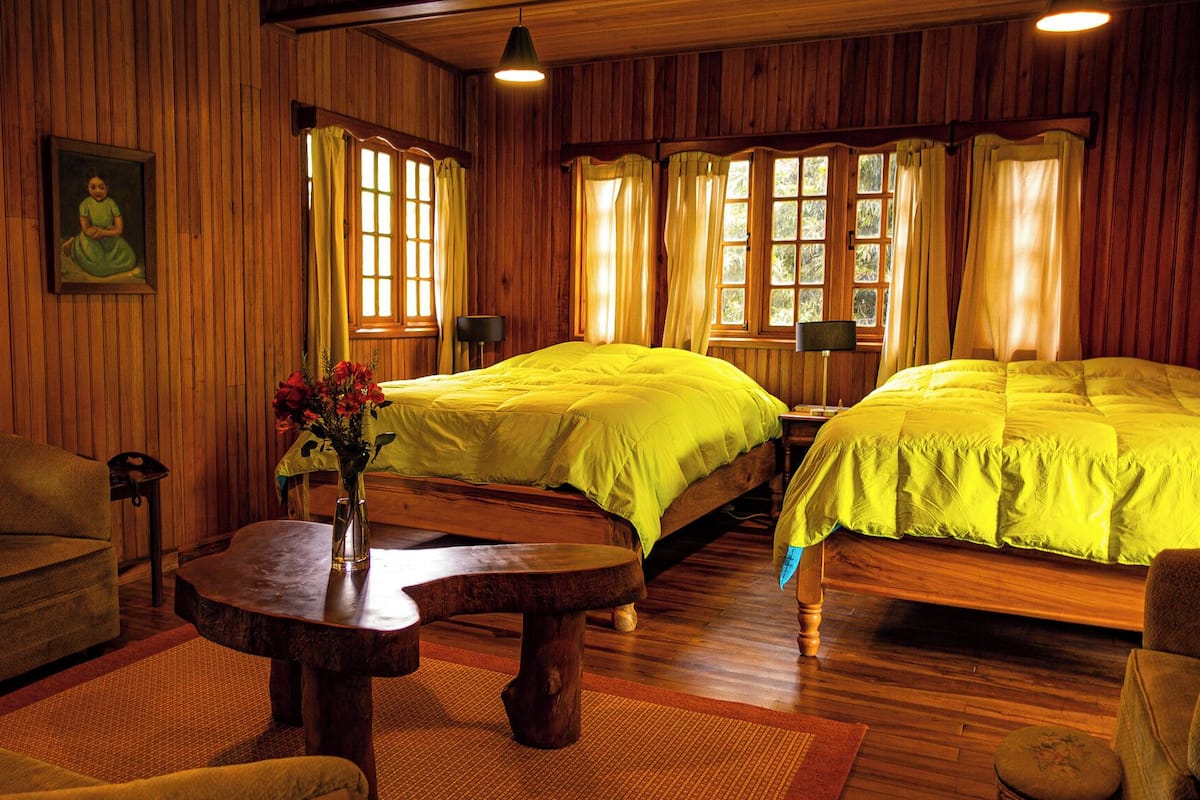 A room with wooden walls, two beds with bright yellow bedding, a wooden table with a vase of flowers, and a chair.