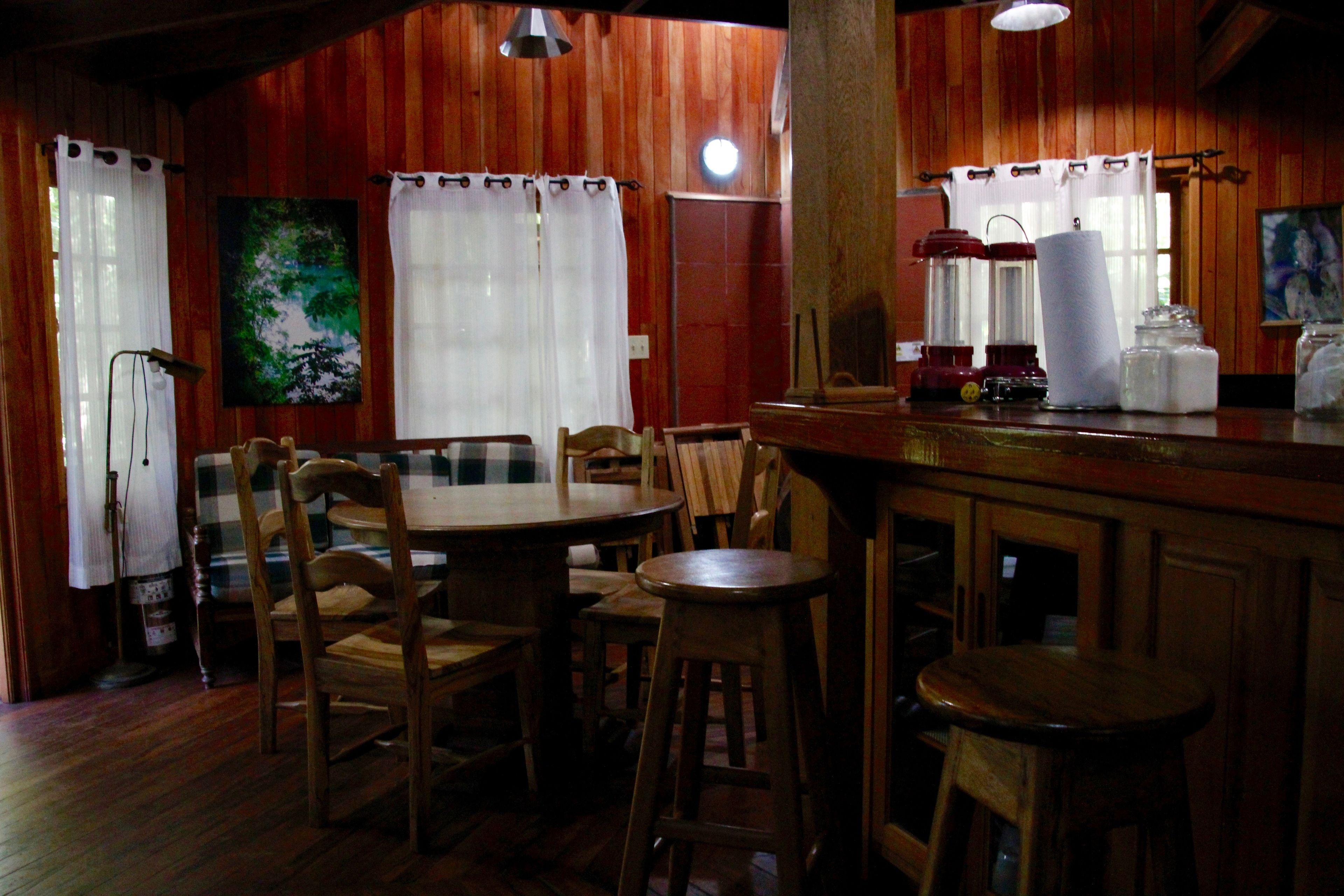 A cozy dining area with wooden walls and a wooden table surrounded by chairs. There are curtains on the windows, a painting on the wall, and a bar area with stools and a counter.