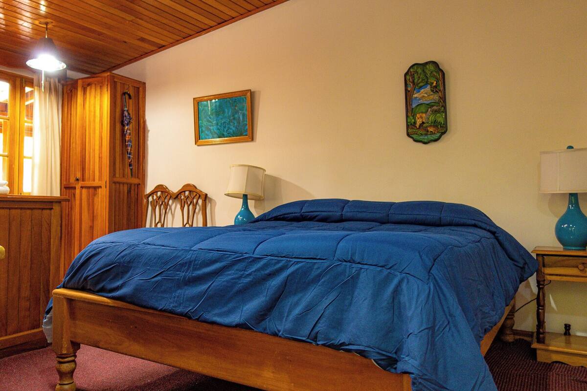 A bedroom with a wooden bed, a wooden wardrobe, a blue lamp, and a painting on the wall.