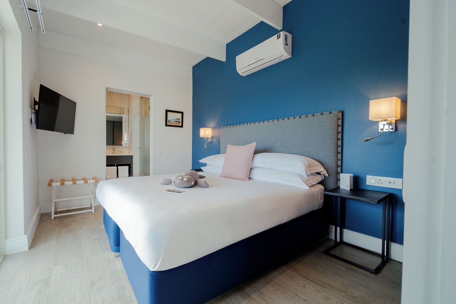 Una habitación de hotel moderna con una cama grande, un televisor de pantalla plana, una mesita de noche con lámpara y un equipo de aire acondicionado.