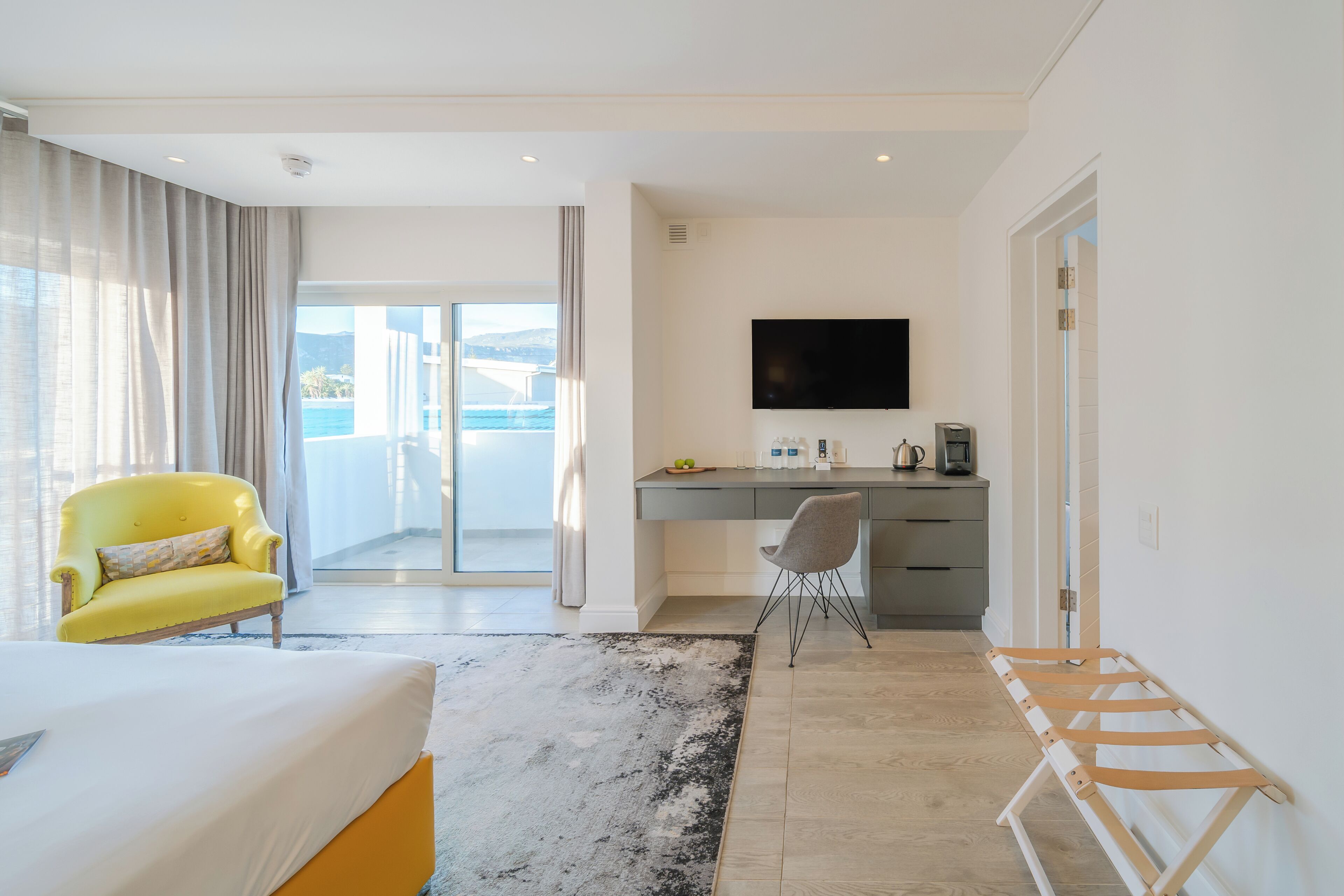 A modern hotel room with a bed, a desk, a chair, a TV, and a view of the sea.