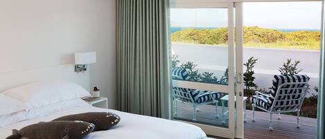A hotel room with a bed, a nightstand, a lamp, a window with a view of the outdoors, and a balcony with chairs.