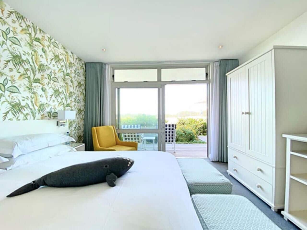 A hotel room with a large bed, a yellow chair, a wardrobe, and a view of the outdoors through a glass door.