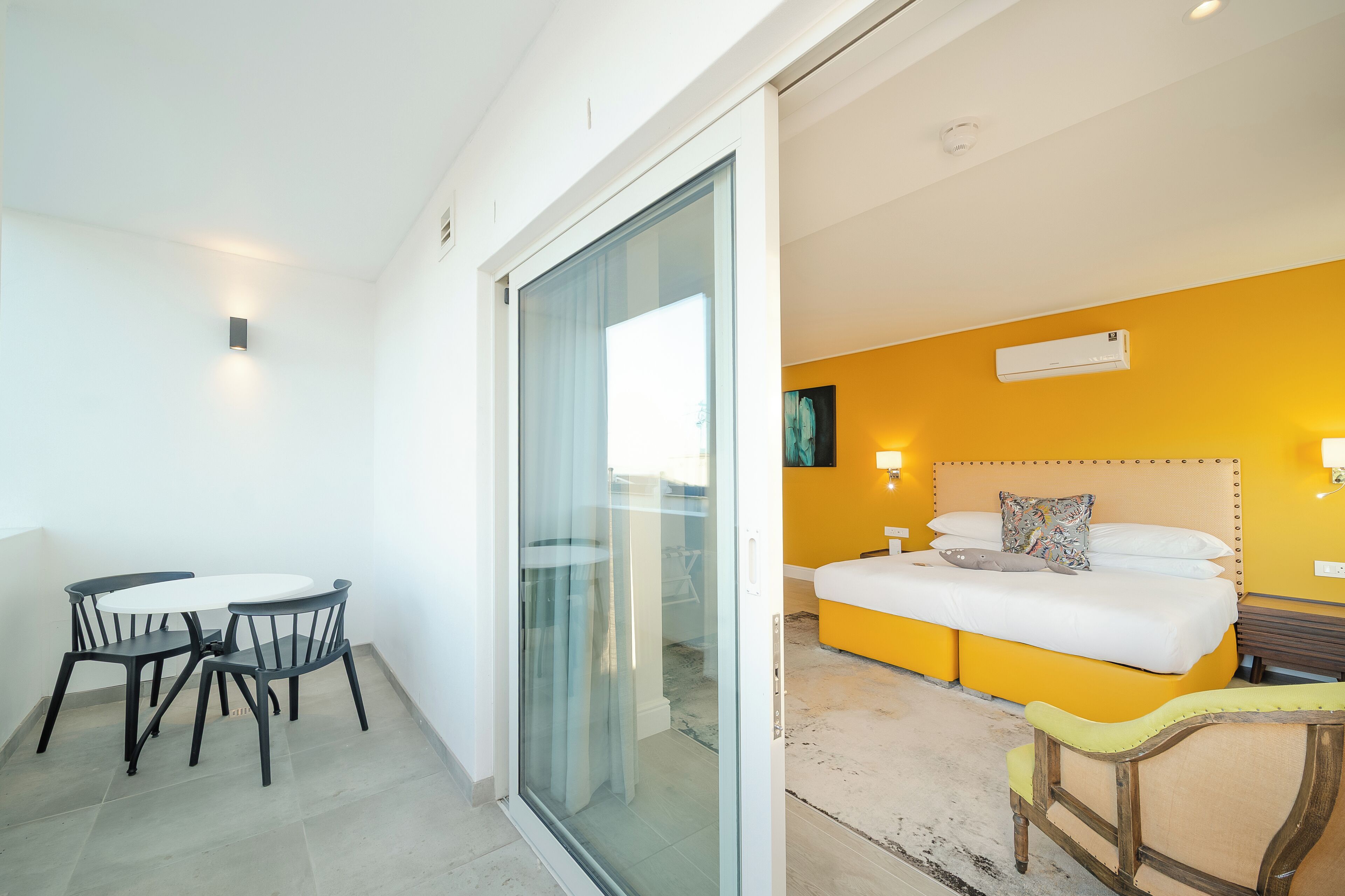 Habitación de hotel con cama, mesitas de noche, una pequeña mesa con sillas y un ventanal corredero que da a un balcón.