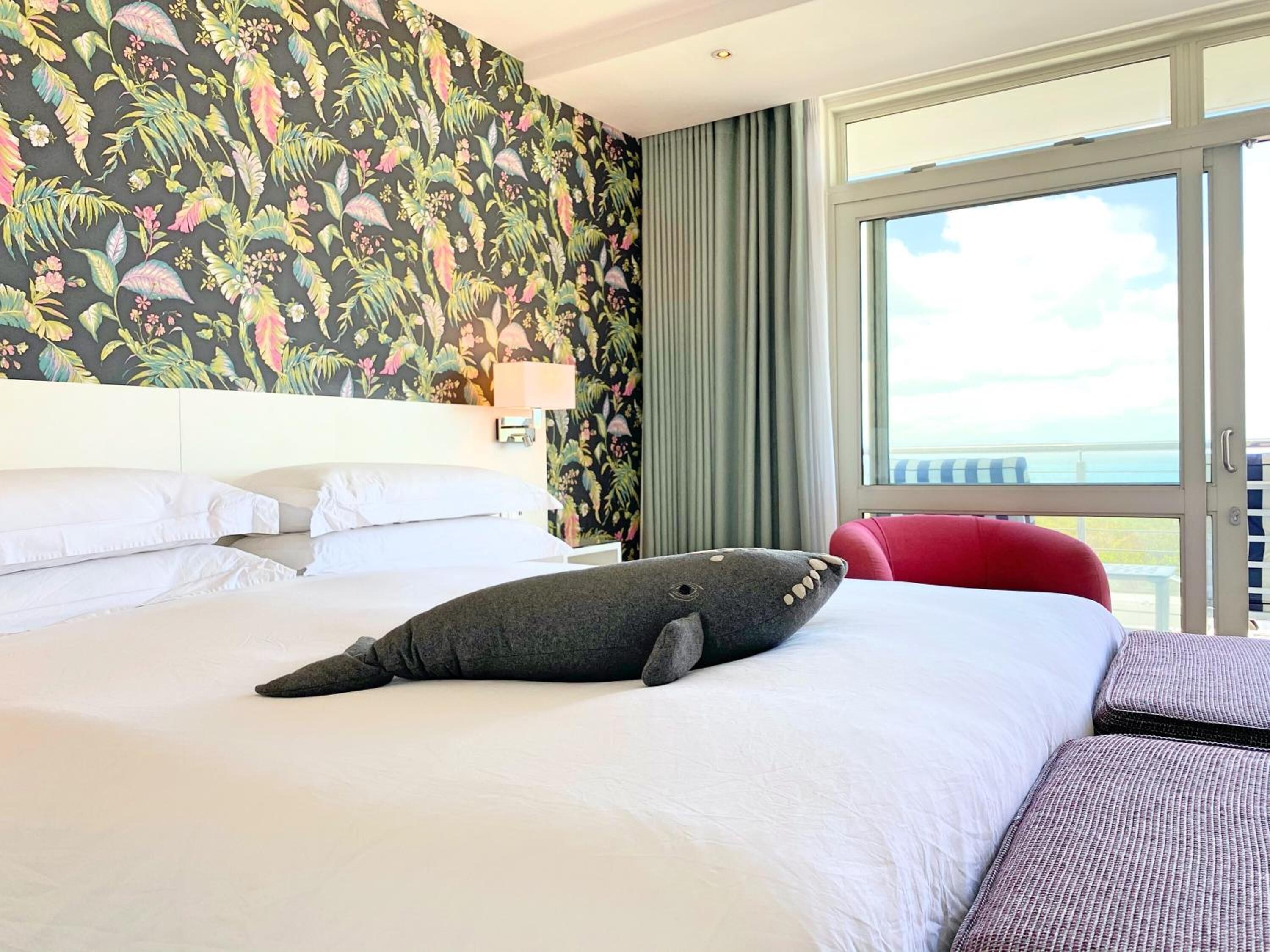 A hotel room with a large bed, a view of the sea through a window, and a tropical floral wallpaper.