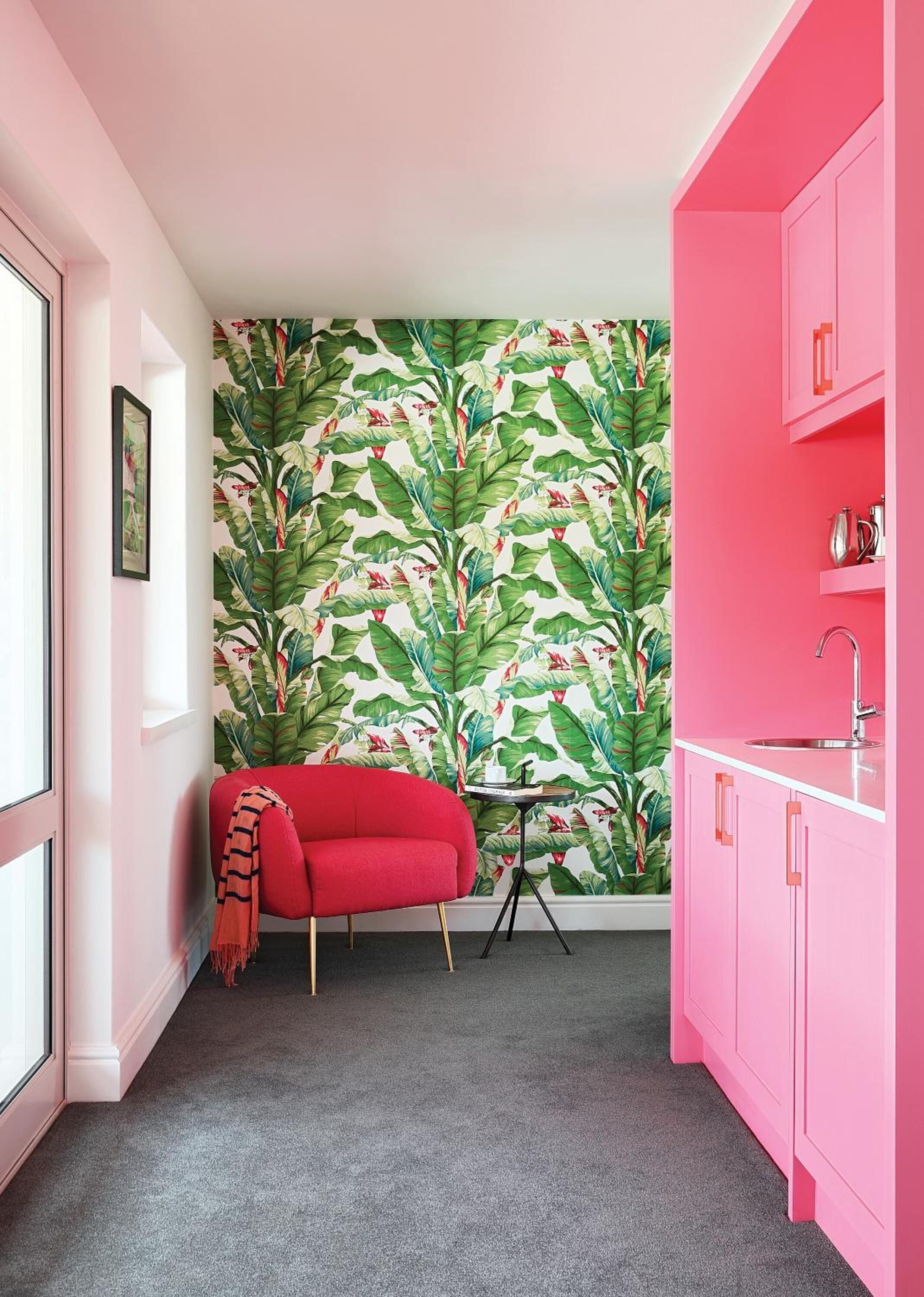 A room with a vibrant pink color scheme, featuring a red armchair, a small round table, and a wall with a tropical leaf pattern.
