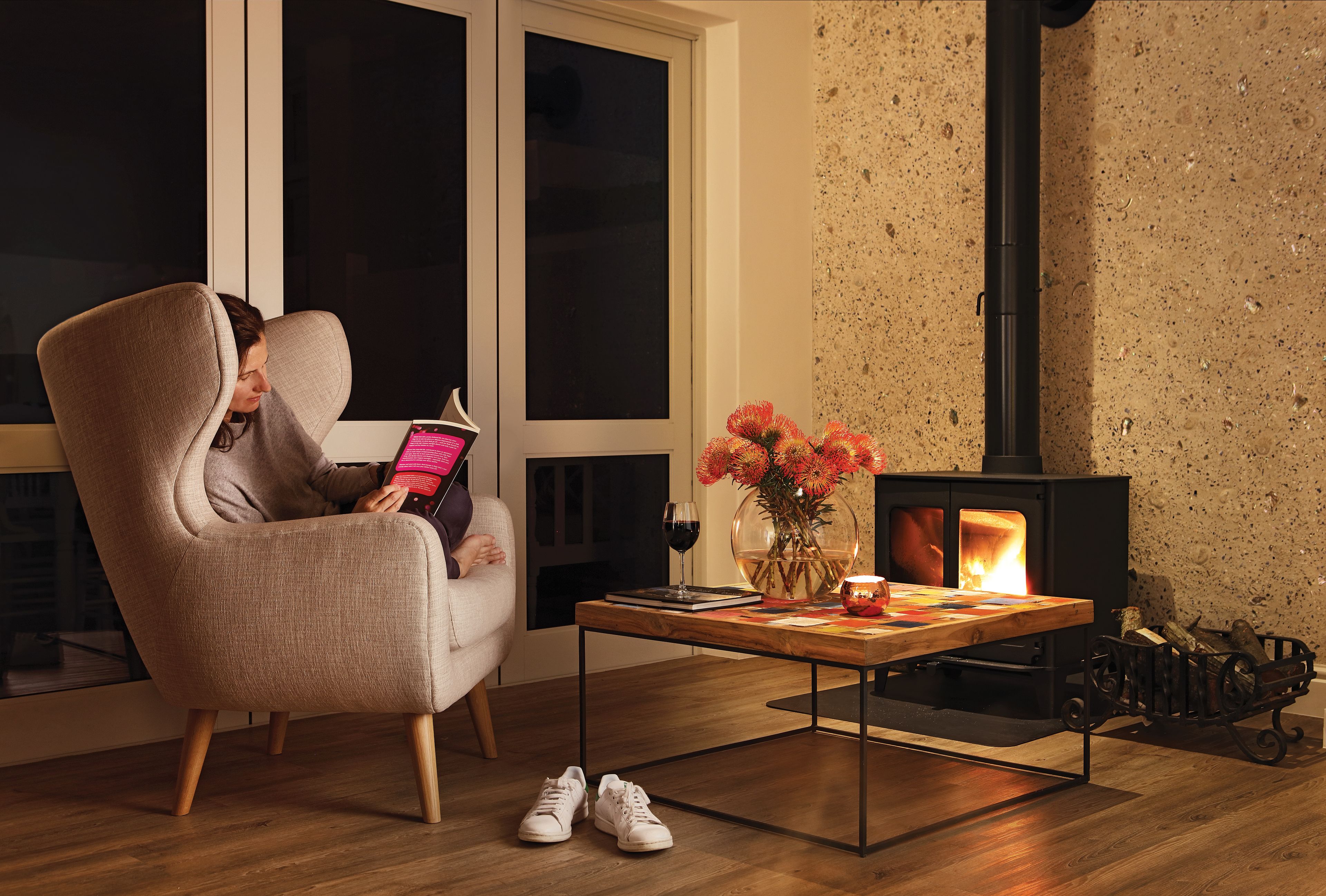 A cozy living room with a person reading a book, a wooden table with a vase of flowers, and a fireplace.