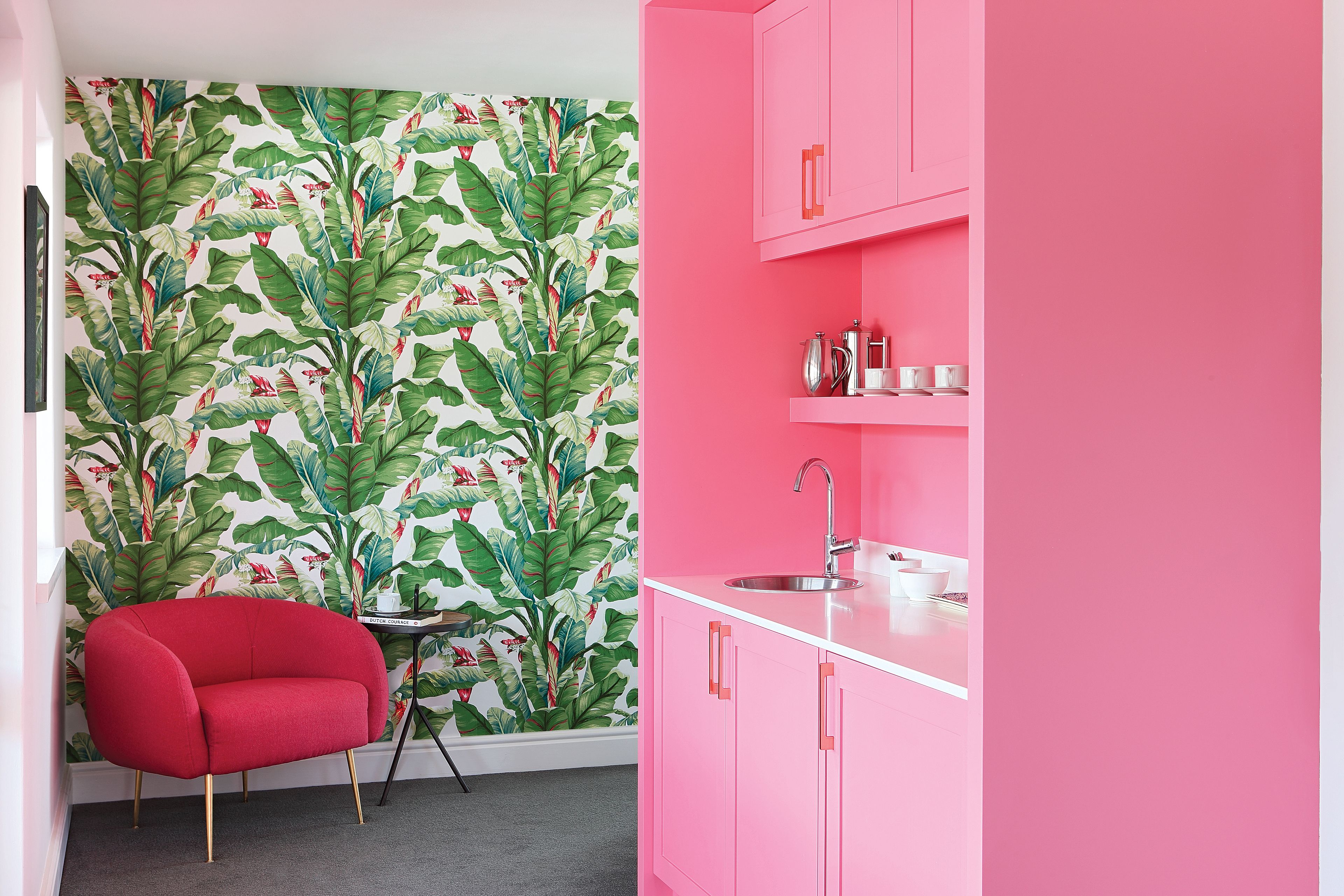 Une cuisine avec des placards roses, une chaise rouge et un mur orné d’un motif de feuilles tropicales.