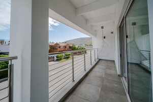 Un balcón con barandilla de metal, puerta de cristal y vistas a casas y montañas al fondo.