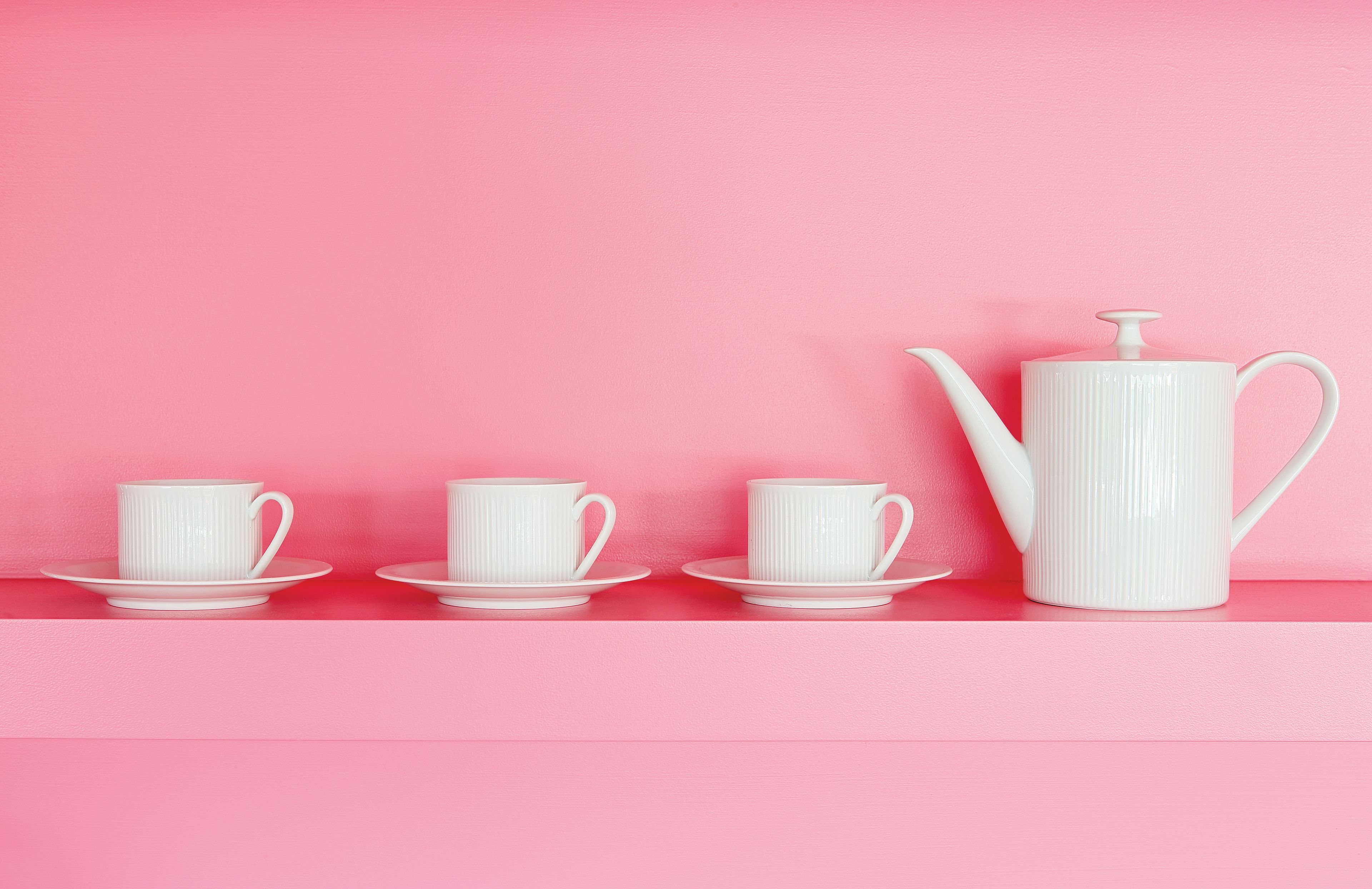 A pink shelf with three white cups and saucers, and a white teapot.