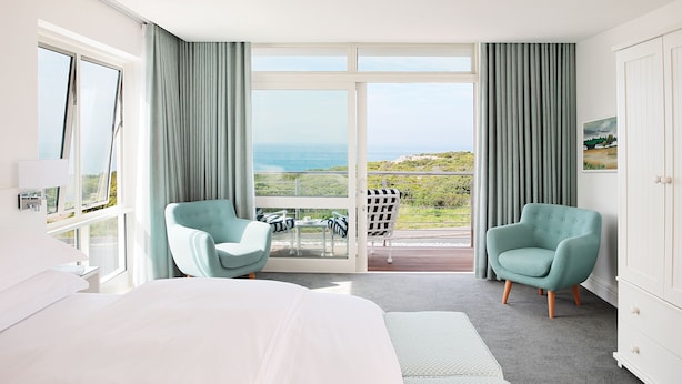 A hotel room with a large bed, two armchairs, a balcony with a view of the sea, and a white wardrobe.