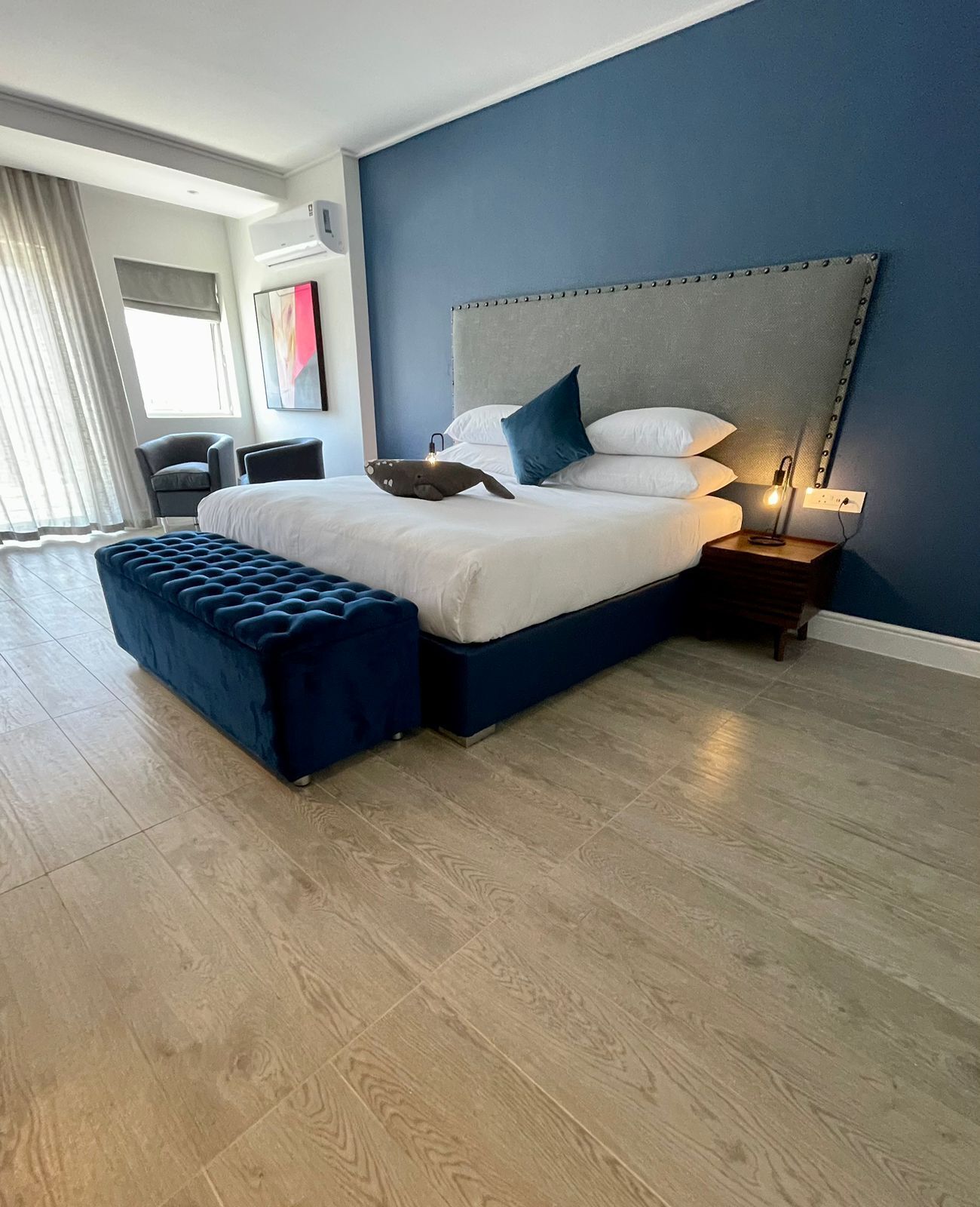 A modern bedroom with a large bed, a bench, a nightstand, and a wall-mounted air conditioner.