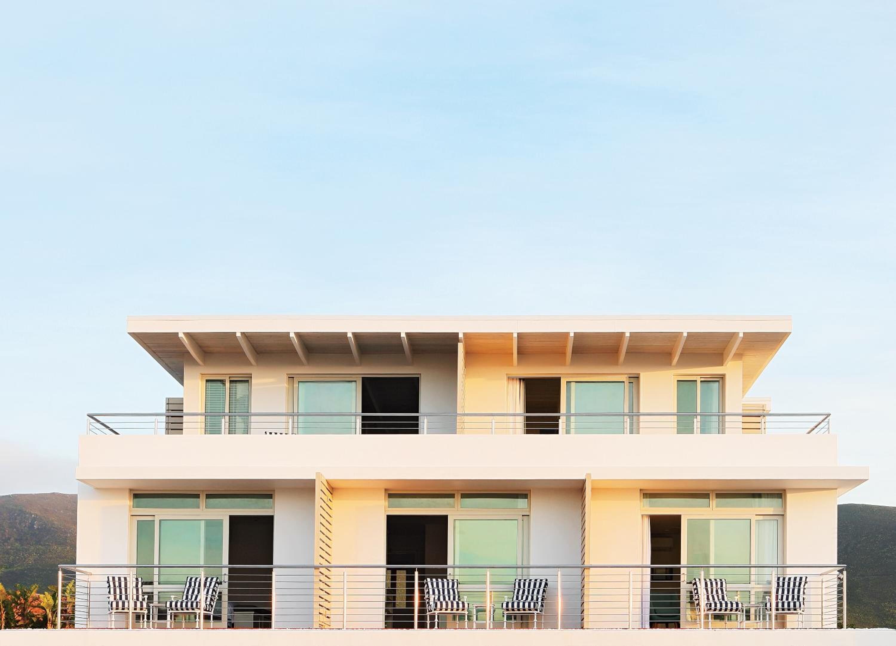 A modern two-story building with a flat roof, large windows, and a balcony with outdoor seating.