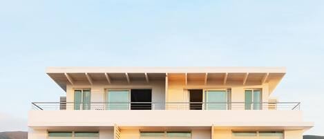 A modern two-story building with a flat roof, large windows, and a balcony with outdoor seating.