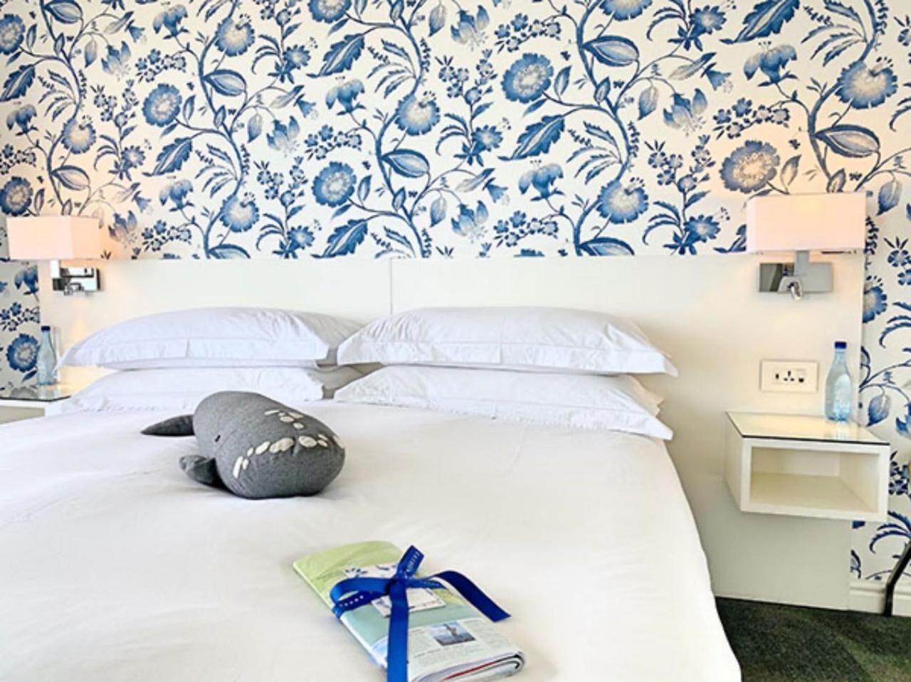 A hotel room with a floral wallpaper, a bed with white linens, a grey pillow, and a magazine on the bed.