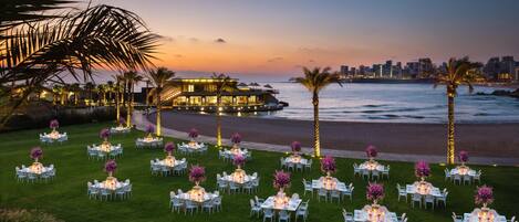 An outdoor event space with multiple round tables set up on a lawn, overlooking a body of water and a city skyline at dusk.