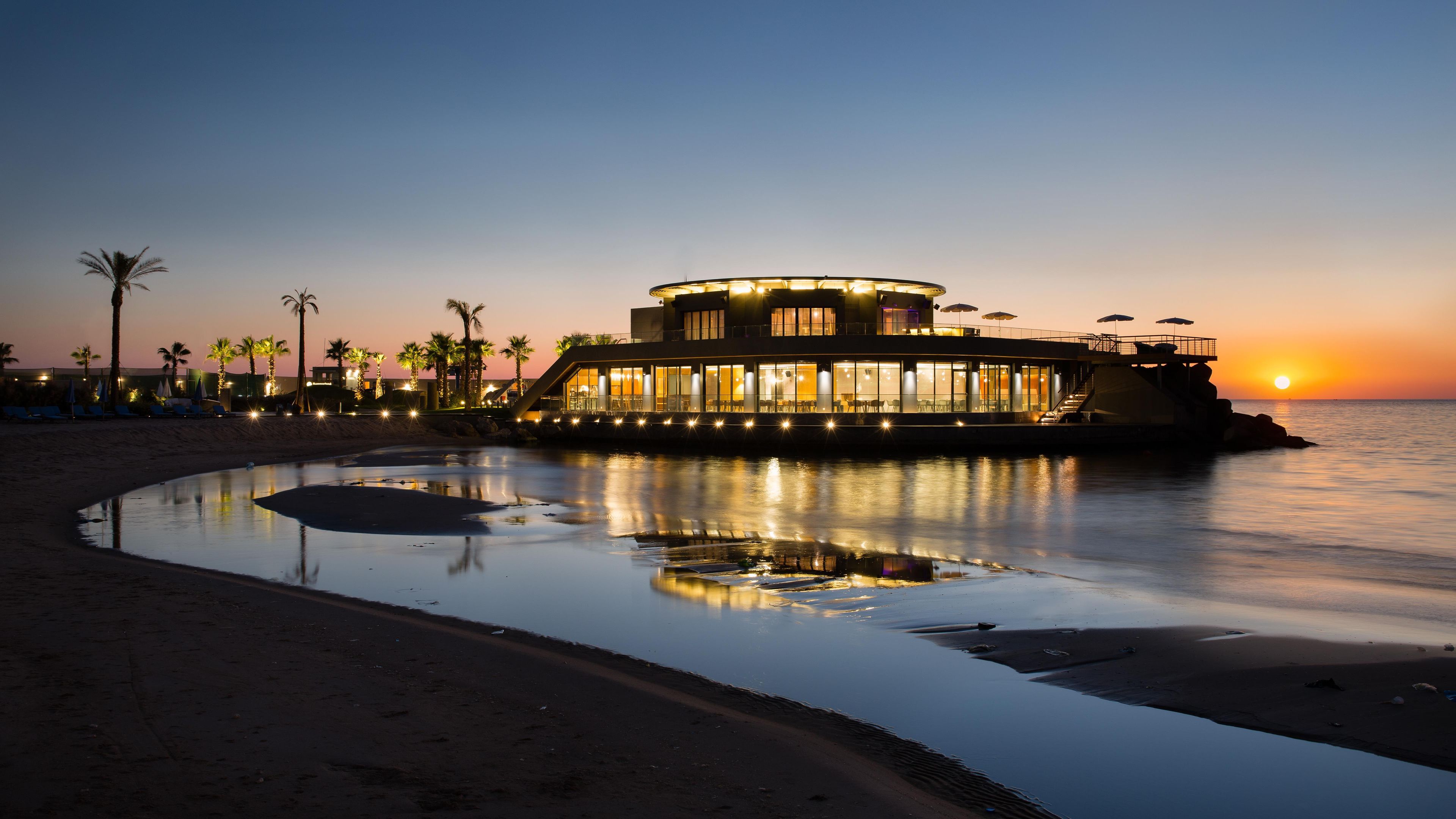 A waterfront building with a circular structure, a pool, and palm trees.