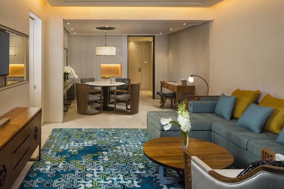 Una habitación de hotel moderna con una televisión de pantalla plana, un sofá con cojines azules y amarillos, una alfombra estampada y un área de comedor con mesa y sillas.