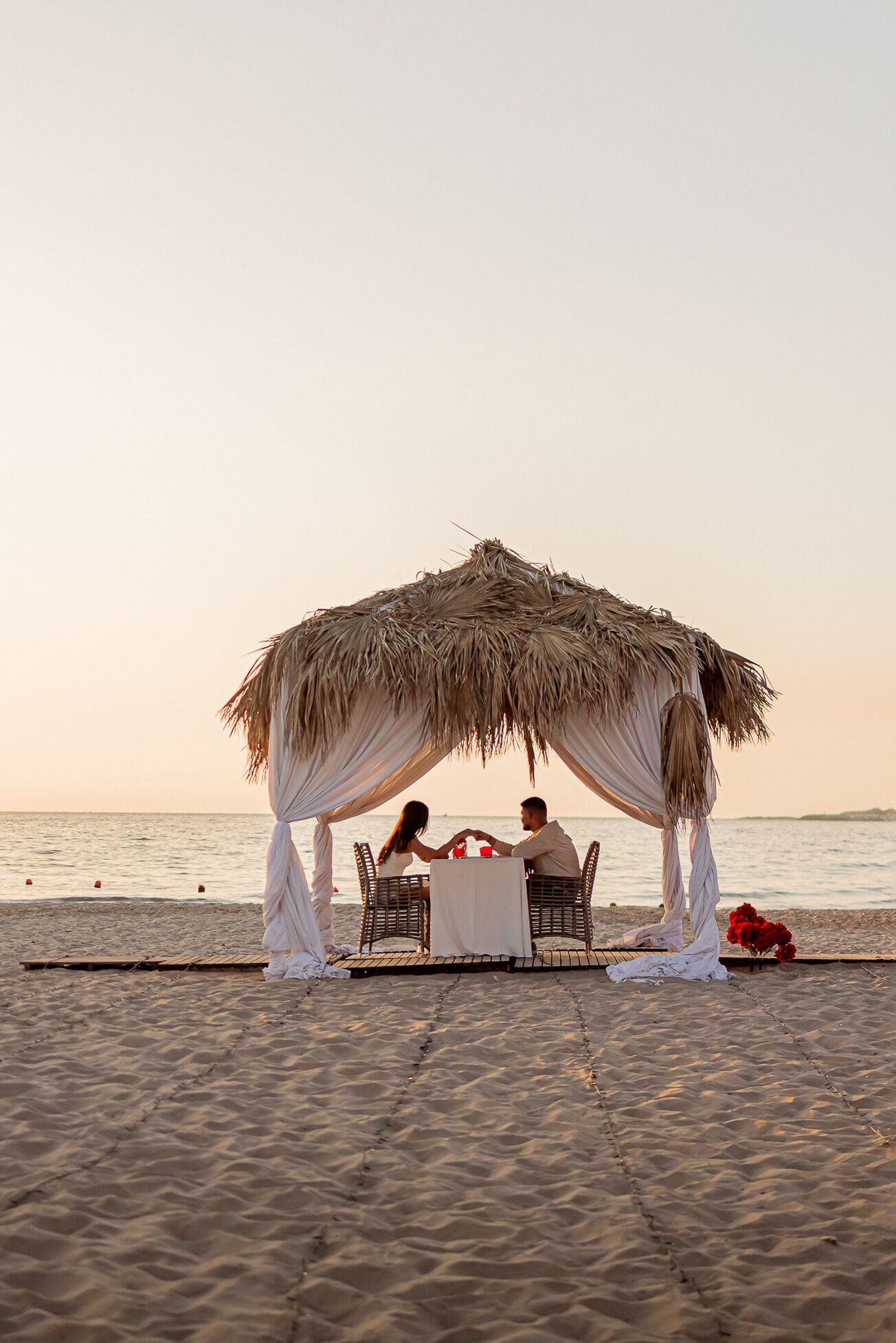 Un montaje para comer frente al mar, con un toldo de paja, sillas de mimbre y una mesa cubierta con mantel blanco.