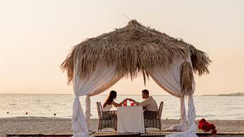 Un montaje para comer frente al mar, con un toldo de paja, sillas de mimbre y una mesa cubierta con mantel blanco.