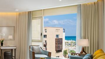 A hotel room with a large window offering a view of the sea and a building across the water. There is a desk with a lamp, a chair, and a sofa with cushions.