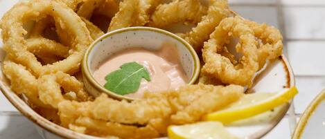 Un bol de calamars frits, accompagné de quartiers de citron et d’une sauce pour tremper, posé sur une table carrelée.