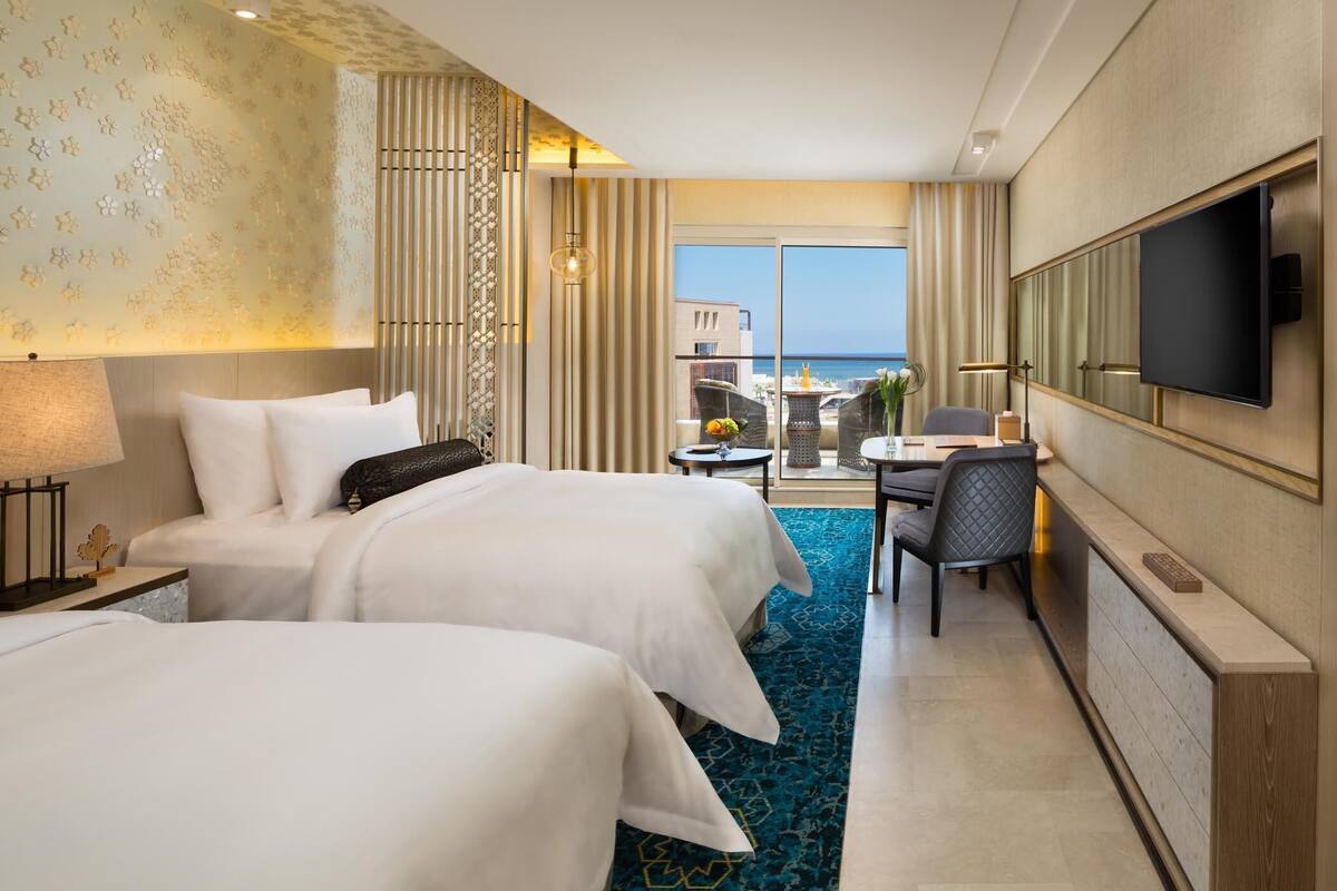 Habitación de hotel con dos camas, un escritorio, televisor y vista al mar.