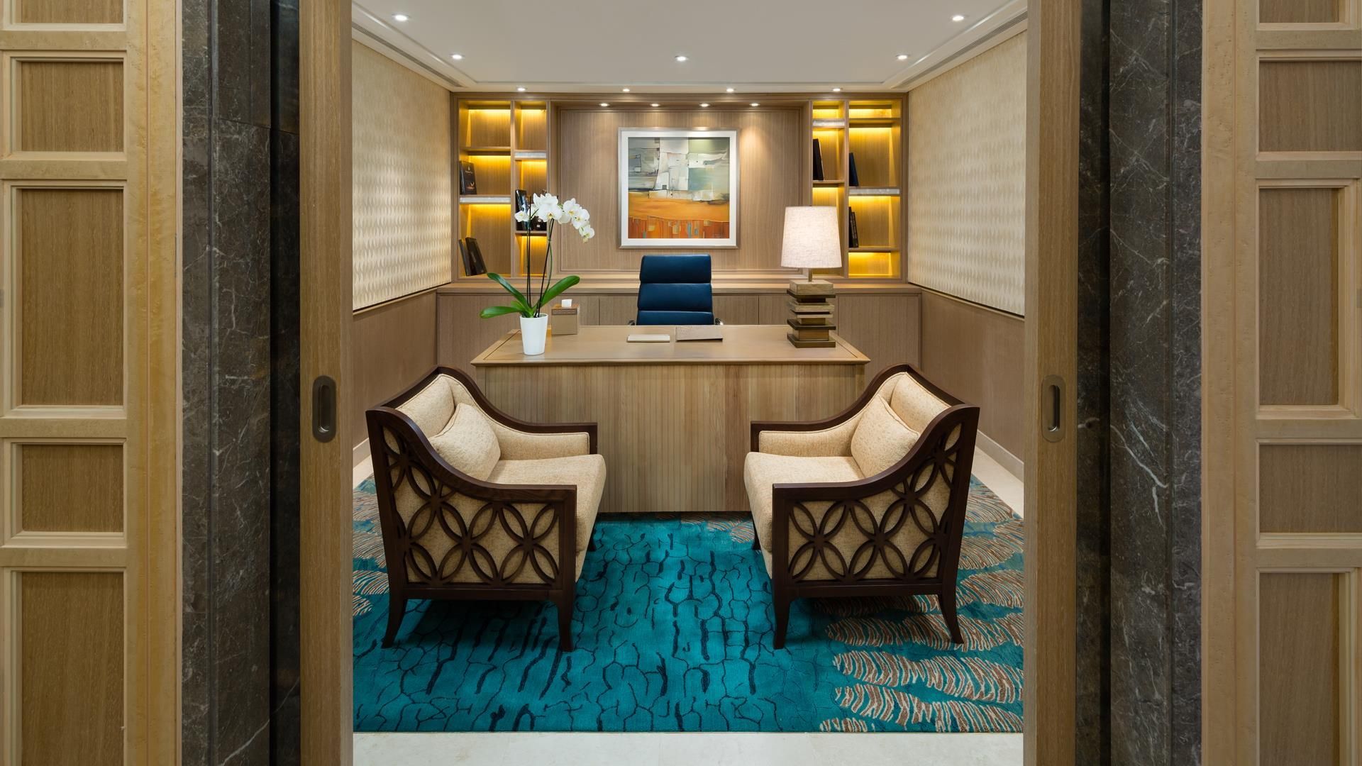 A hotel room with a desk, two chairs, a blue sofa, a patterned rug, and a framed painting on the wall.