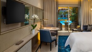 Habitación de hotel con cama, escritorio con silla, televisor montado en la pared, pequeña mesa con una silla, planta en maceta y vista a una piscina por la ventana.