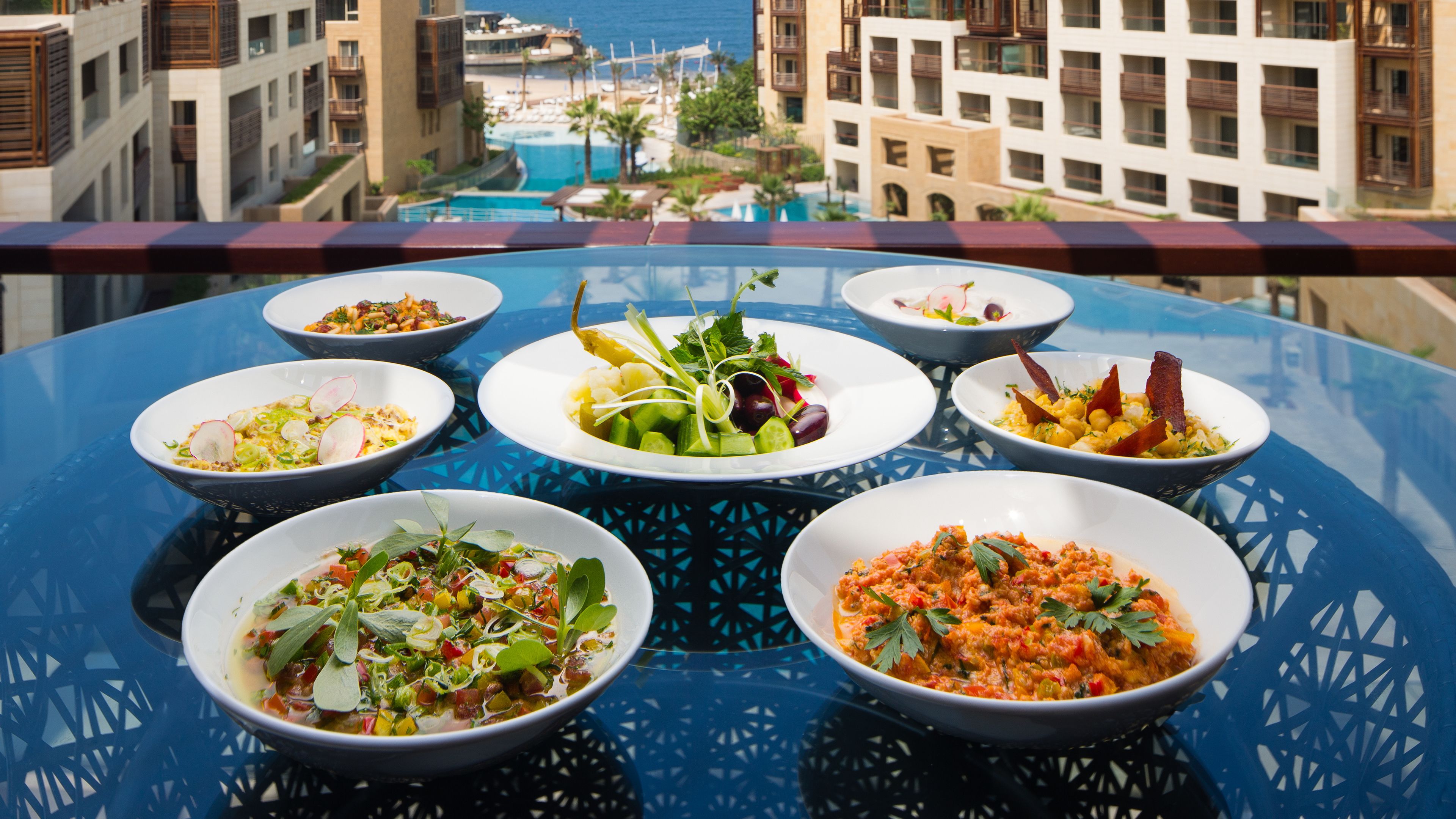 A table with various dishes on a balcony overlooking a pool and buildings.