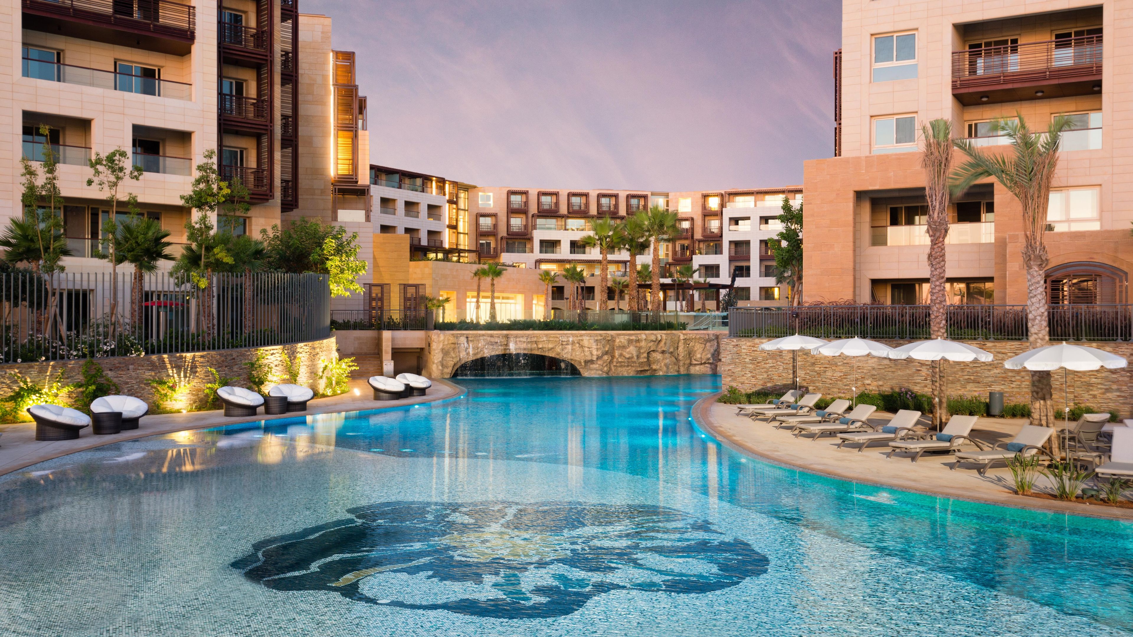 A hotel pool area with a circular water feature, lounge chairs, and a stone bridge.