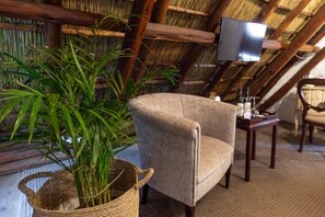 A cozy room with a thatched ceiling, a comfortable armchair, a small table with bottles, and a television mounted on the wall.