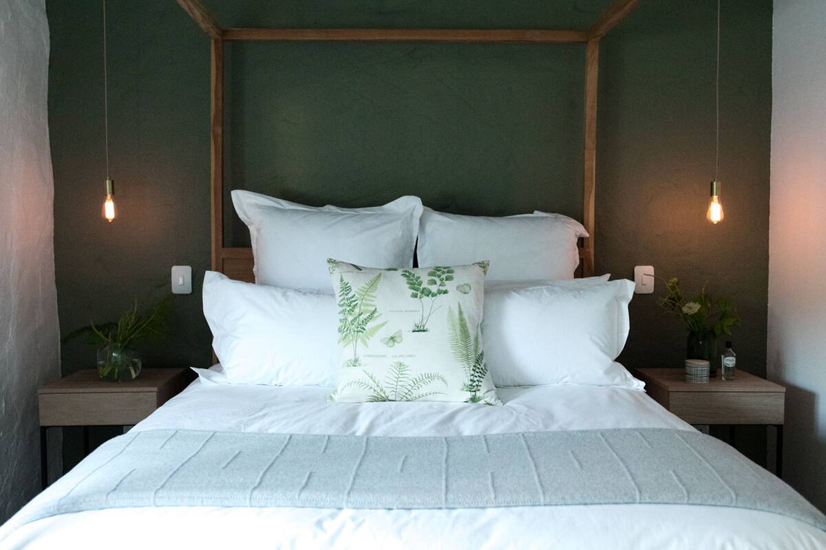 A neatly made bed with white linens and a botanical pillow, flanked by two bedside tables with small plants and a bottle.
