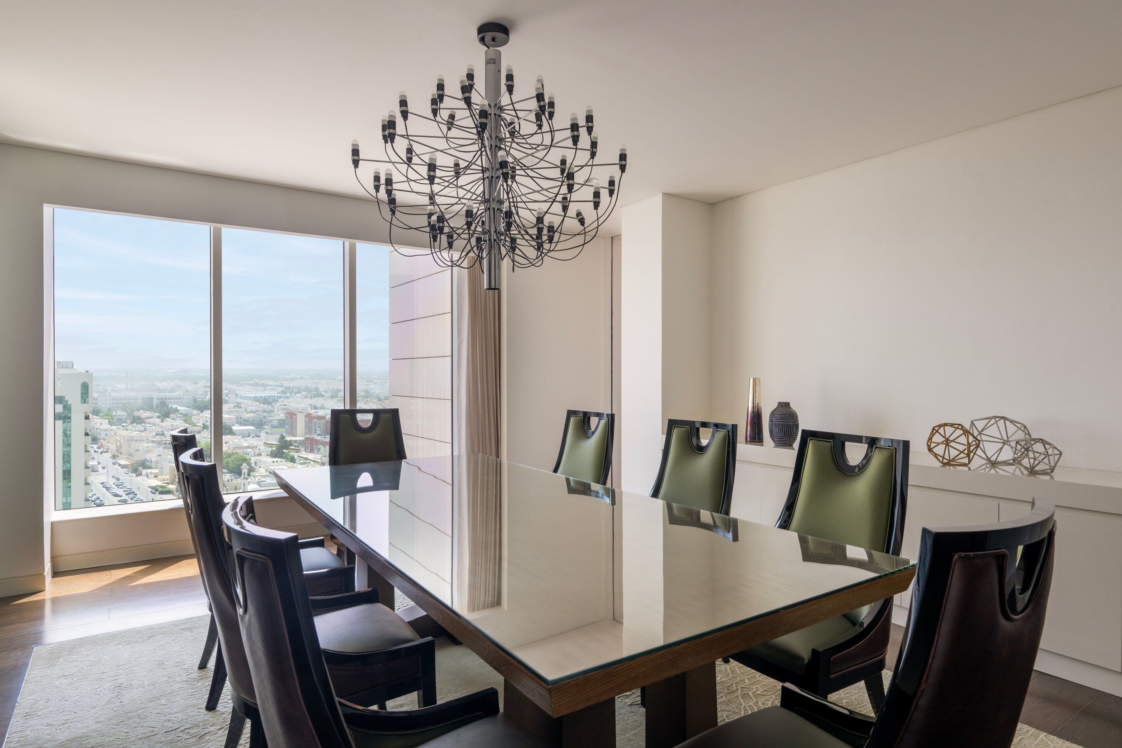 A modern dining room with a glass table and chairs, a unique chandelier, and a large window offering a city view.