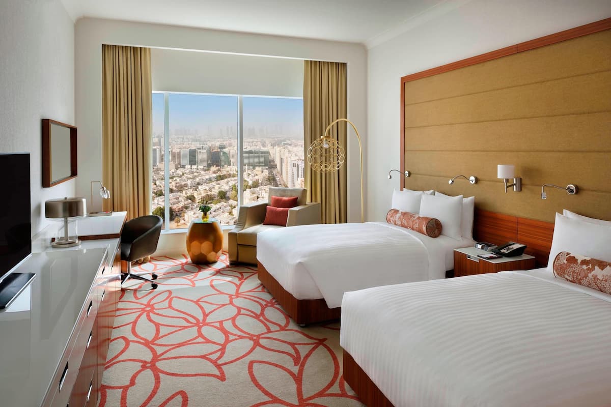 A hotel room with two beds, a desk, a chair, a TV, and a large window with a city view.