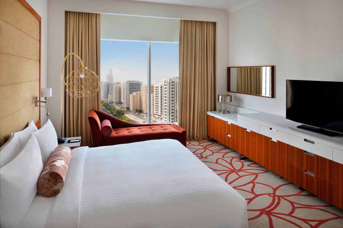 A hotel room with a large bed, a TV, a desk, and a view of the city.