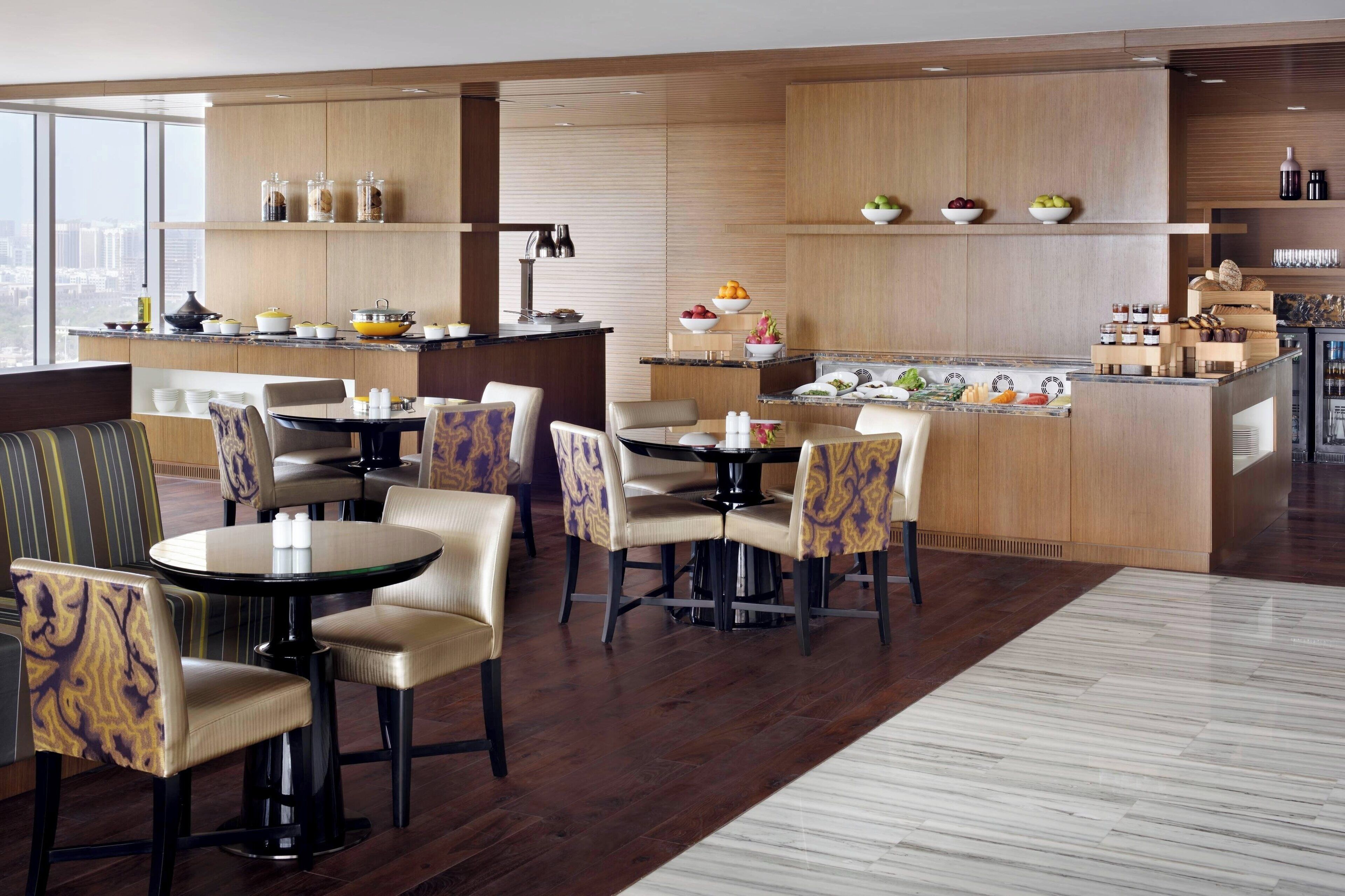 A modern dining area with a central island, multiple seating arrangements, and a kitchen area with wooden cabinets and a view of the city.