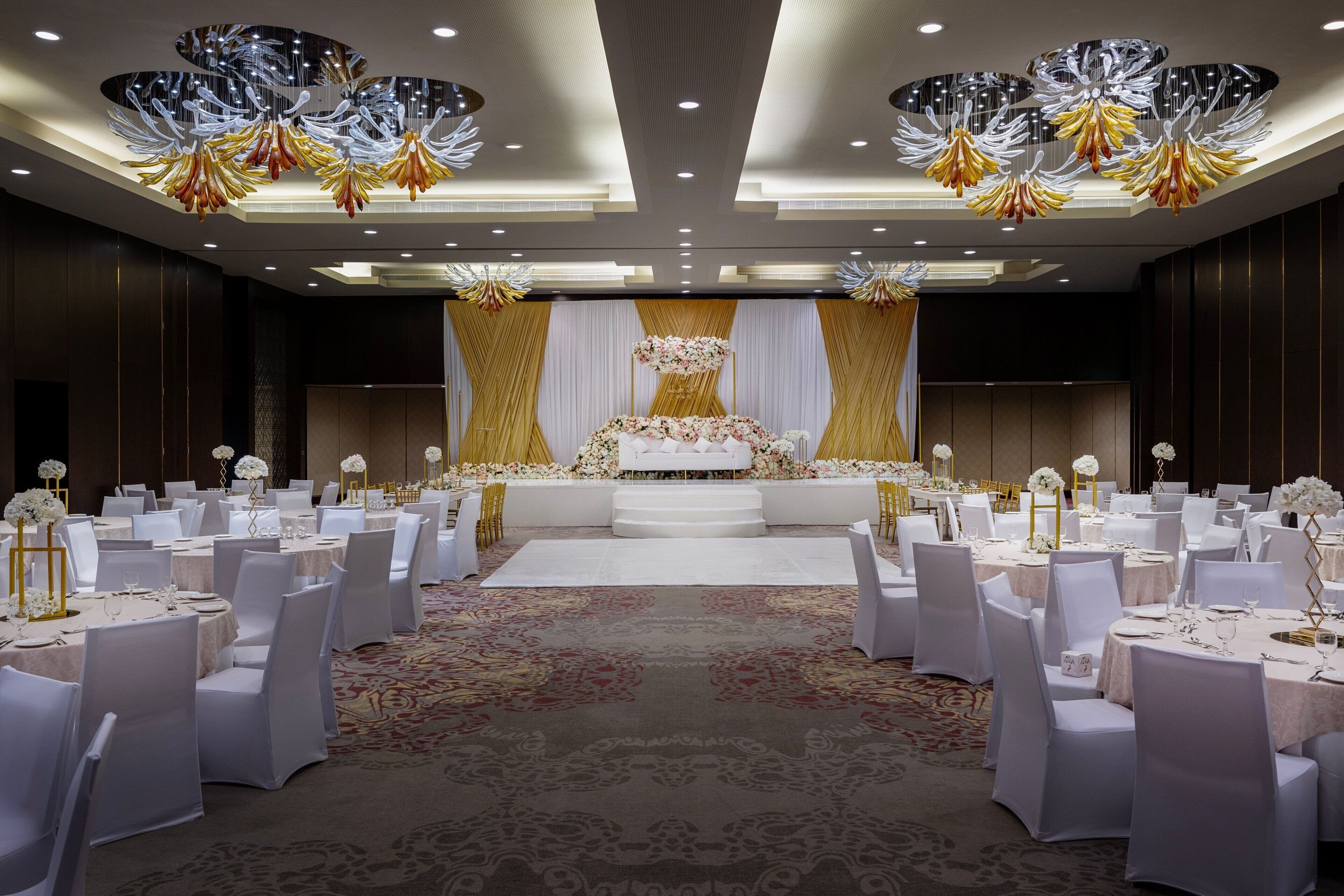 Une salle de banquet avec des tables rondes, des chaises blanches et une scène centrale agrémentée de décorations florales.