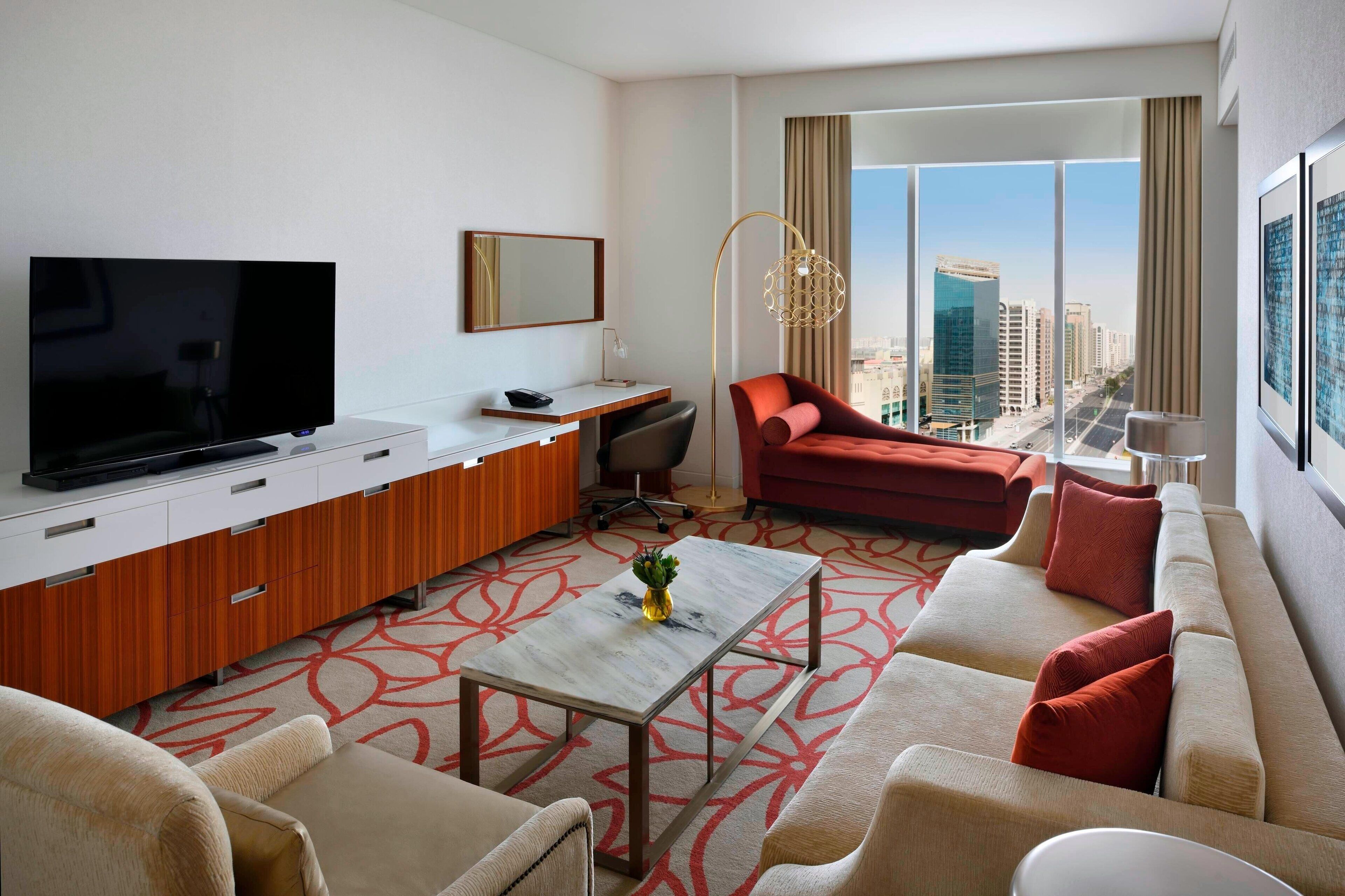 A modern hotel room with a large flat-screen TV, a sofa, a red armchair, a small table with a vase, and a city view through large windows.