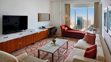 A modern hotel room with a large flat-screen TV, a sofa, a red armchair, a small table with a vase, and a city view through large windows.