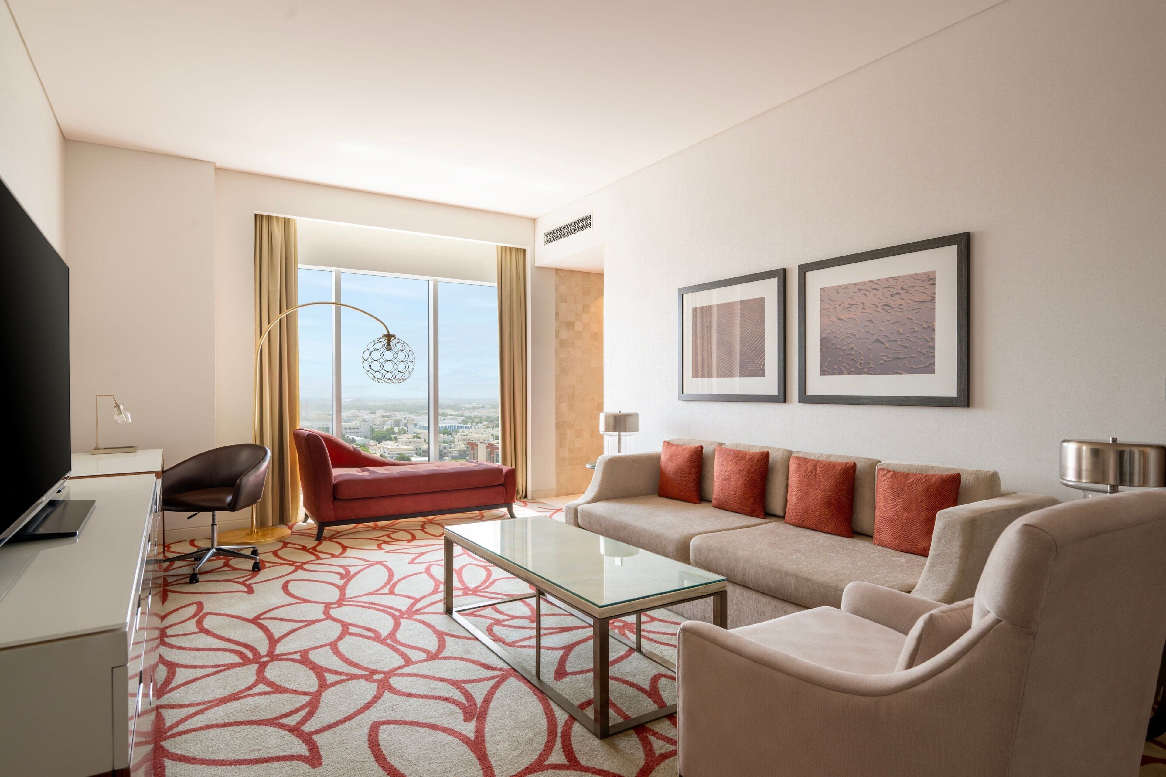 A modern hotel room with a large window, a sofa, a red armchair, a glass coffee table, and a flat-screen TV.