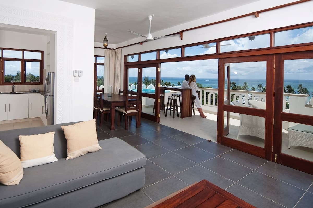 A modern living room with a grey sofa, a dining area with a table and chairs, and a balcony with a view of the ocean.