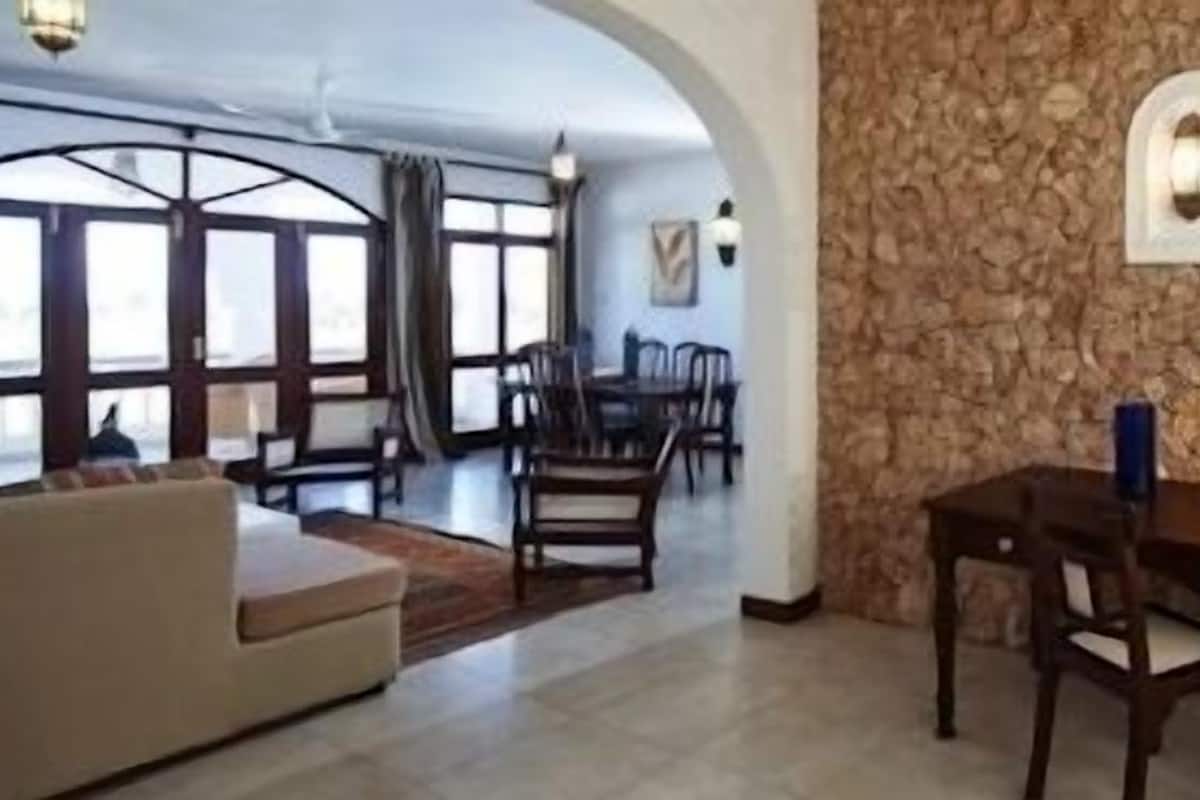A spacious living area with a stone wall, a sofa, a dining table with chairs, and large windows.