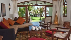 Un salon chaleureux avec un canapé, des fauteuils et une table basse. On peut admirer un jardin avec des palmiers, et un coin repas avec une table dressée pour deux personnes.
