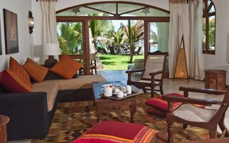 Ein gemütliches Wohnzimmer mit einer Couch, Sesseln und einem Couchtisch. Blick auf einen Garten mit Palmen und eine Essecke mit Tisch für zwei Personen.