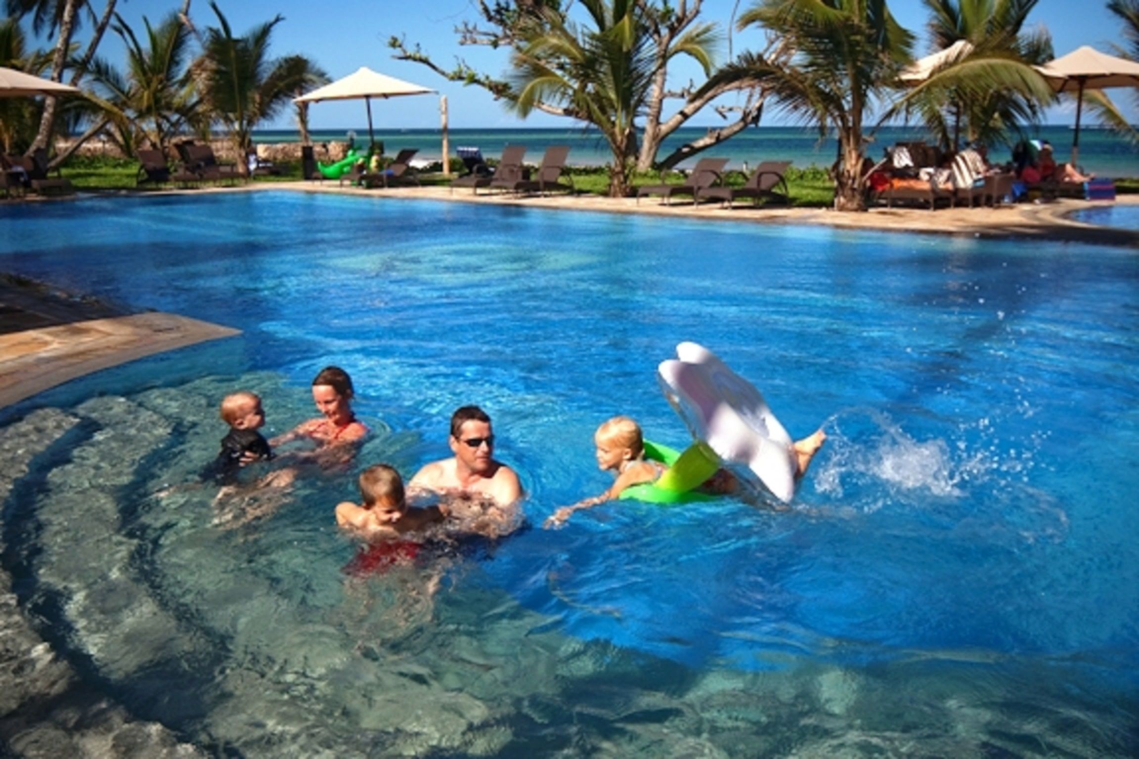 A swimming pool with clear blue water, surrounded by lounge chairs and umbrellas, with a view of the ocean in the background.