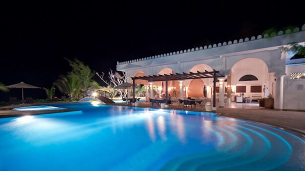 Un espace piscine nocturne doté d’un coin salon couvert et, en arrière-plan, d’un bâtiment orné d’arches.