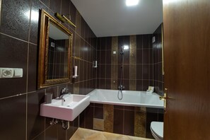 A bathroom with a white sink, a large mirror, a bathtub, and dark patterned tiles. - Kostelski Buk Hotel (Bihac)