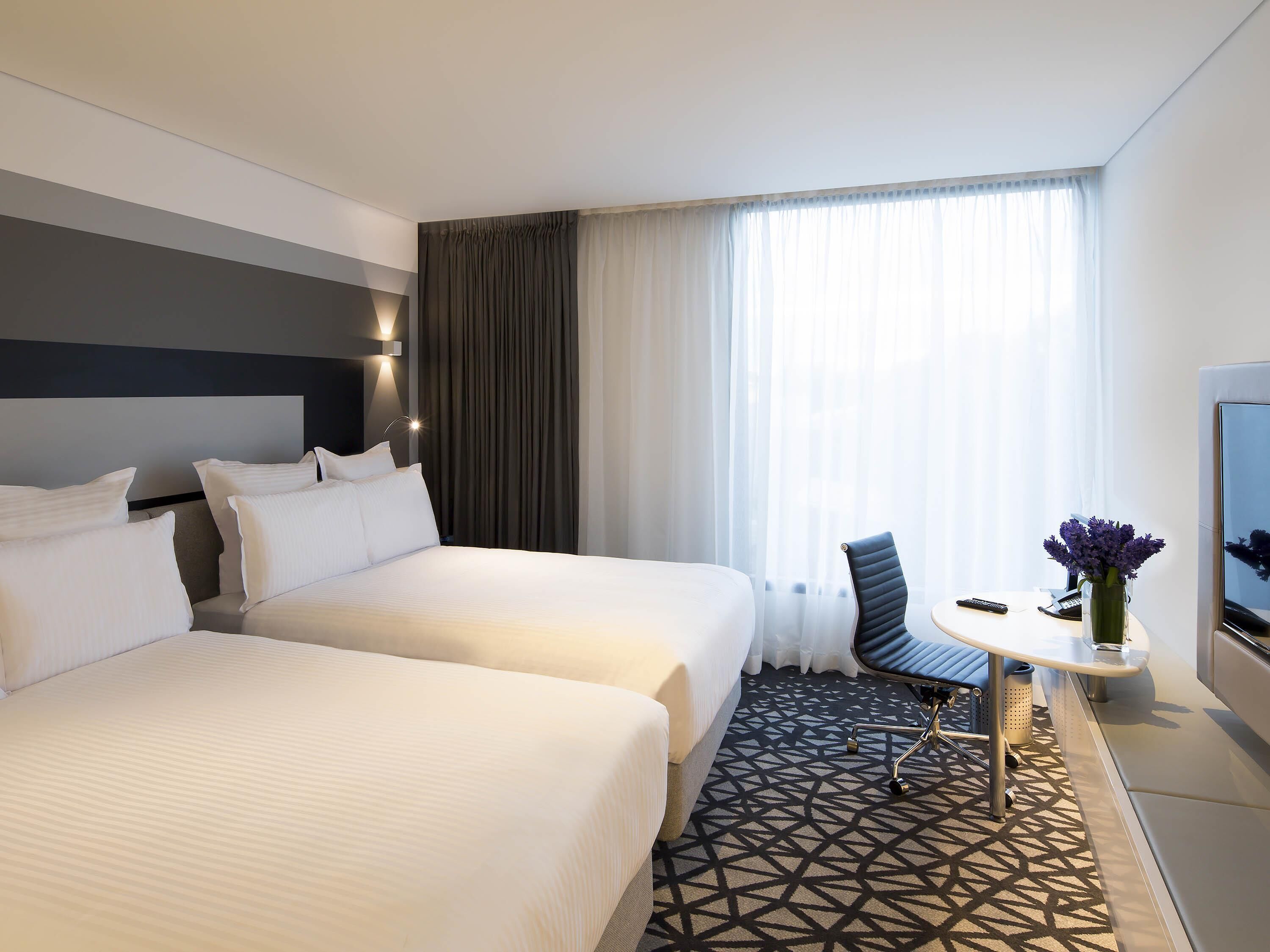 A hotel room with two beds, a desk with a chair, a TV, and a window with curtains.