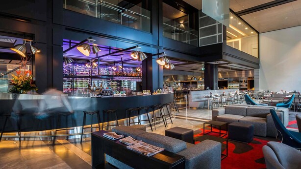 A modern bar area with a high ceiling, bar stools, and a seating area with sofas and tables. - Pullman Sydney Airport (Mascot)